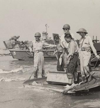 Black and white photo of US military on boats and ships in the background