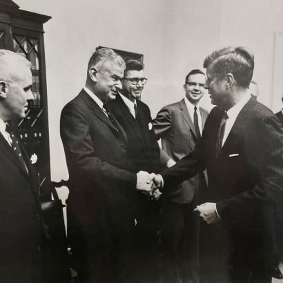 Black and white photo of 5 men in a room. Norman Obbard shaking hands with President John F. Kennedy.