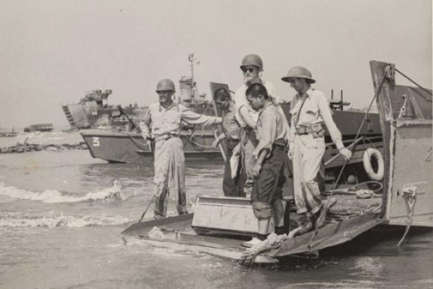 Black and white photo of US military on boats and ships in the background