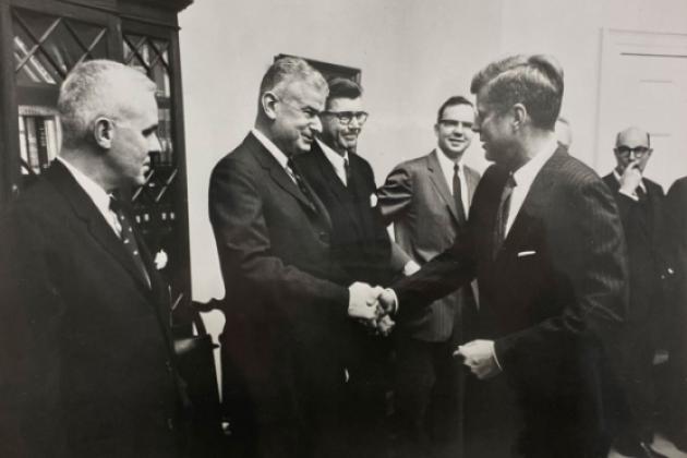 Black and white photo of 5 men in a room. Norman Obbard shaking hands with President John F. Kennedy.