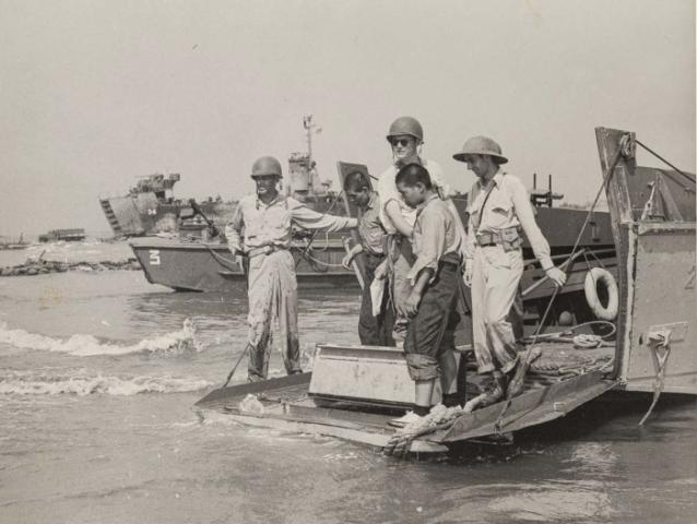 Black and white photo of US military on boats and ships in the background